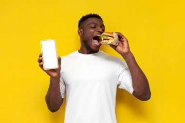 Beyaz tişörtlü Afro-Amerikan adam büyük bir burger tutuyor ve sarı izole edilmiş arka planda boş akıllı telefon ekranı gösteriyor, genç adam fast food yiyor ve telefon reklamı yapıyor, uzayı kopyalıyor.