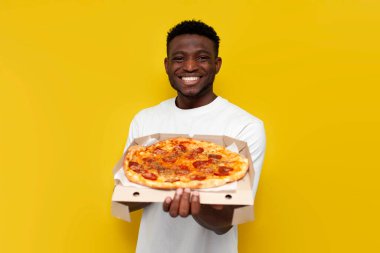 Beyaz tişörtlü neşeli Afro-Amerikalı adam bir kutu pizza tutuyor ve sarı izole edilmiş arka planda gülümsüyor. Adam fast food reklamları yapıyor.