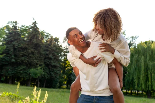 アフリカ系アメリカ人の幸せなカップルが夏に公園を歩いて笑顔で 男は彼の背中に女性を運び 自然の中で抱擁 自由と関係の概念 — ストック写真