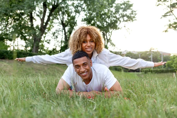 アフリカ系アメリカ人の若い恋人たちが夏に公園の草の上に横になり笑顔で彼女のボーイフレンドの背中に横たわって喜び — ストック写真