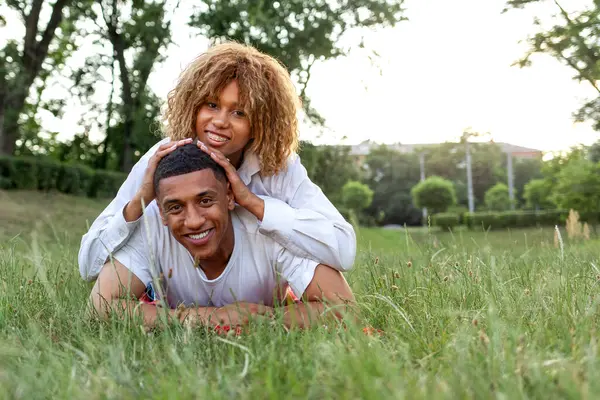 アフリカ系アメリカ人の若い恋人たちが夏に公園の芝生の上に横になって笑顔で彼女のボーイフレンドの背中に横たわって喜び スペースをコピー — ストック写真
