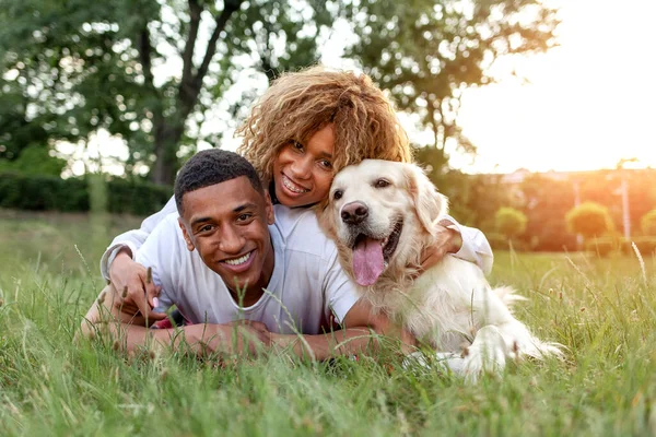 stock image african american guy with girl lie together with golden retriever in the park in summer, young couple walks and plays with dog in nature, happy family with pet