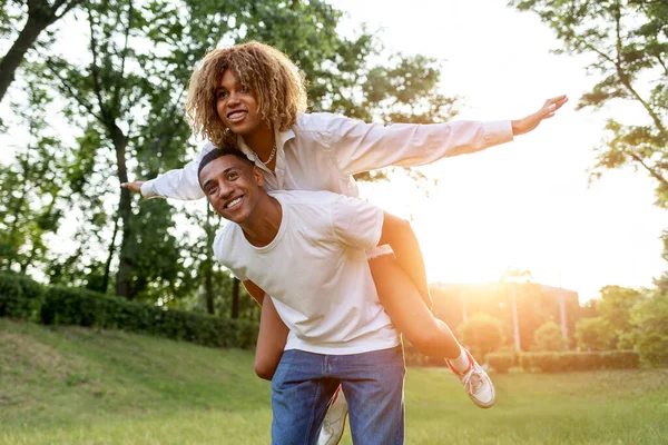 アフリカ系アメリカ人の若いカップルが夏に公園で一緒に走り笑顔で男は彼のバック屋外で彼のガールフレンドを運びます 巻き込ま女の子は前方に飛ぶ 自由と関係の概念 — ストック写真