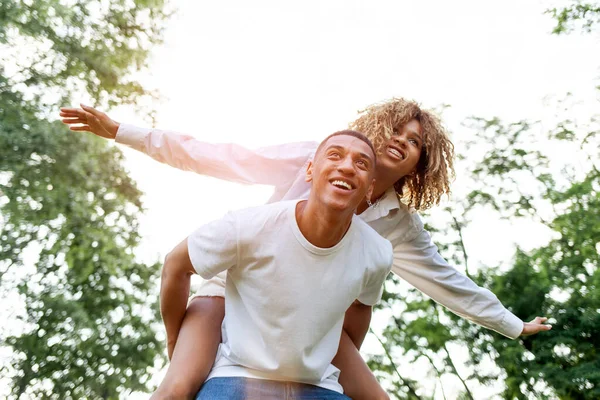 アフリカ系アメリカ人の若いカップルが夏に公園で一緒に走り笑顔で男は彼のバック屋外で彼のガールフレンドを運びます 巻き込ま女の子は前方に飛ぶ 自由と関係の概念 — ストック写真