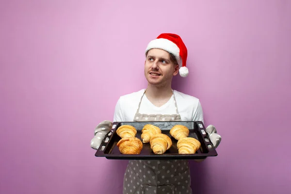 stock image young guy chef in santa hat and apron holds baking sheet with croissants on pink isolated background, male cook in uniform and baking gloves baked pastries for the New Year