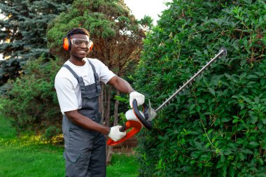 Üniformalı bahçe işçisi çalıları keser, gözlüklü ve kulaklıklı Afro-Amerikan adam bahçede elektrikli aletlerle, budama ağaçlarıyla ve çalılarla çalışır.