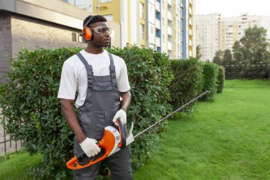 Üniformalı bahçe işçisi çalıları keser, gözlüklü ve kulaklıklı Afro-Amerikan adam bahçede elektrikli aletlerle, budama ağaçlarıyla ve çalılarla çalışır.