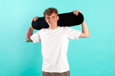 Beyaz tişörtlü, kaykaylı, mavi izole edilmiş arka planda gülümseyen hippi kaykaycı portresi olan genç ve neşeli bir adam.