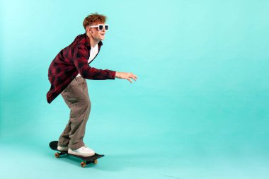 Genç neşeli adam mavi izole arka planda kaykaya biniyor, güneş gözlüklü hippi kaykaycı, kopyalama alanı.