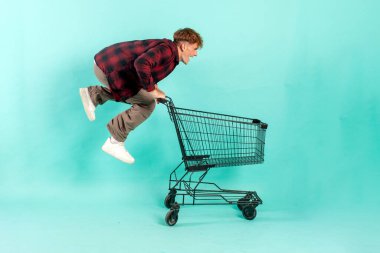 Genç çılgın adam alışveriş arabasıyla mavi izole edilmiş arka planda süpermarketten atlıyor ve seviniyor. Erkek müşteri hızla alışverişe koşuyor.