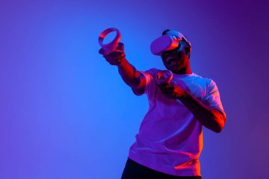 Sanal gerçeklik gözlüklü Afro-Amerikan erkek oyuncu neon ışıklı joystickleri vuruyor ve nişan alıyor, adam modern VR cihazını kullanıyor ve kontrol panelini tutuyor.