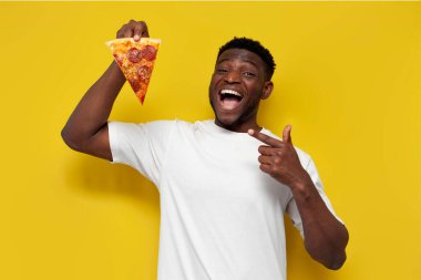 Beyaz tişörtlü neşeli Afro-Amerikan adam bir parça pizza tutuyor ve sarı izole edilmiş arka planda parmağını ona doğrultuyor, adam fast food reklamını yapıyor ve