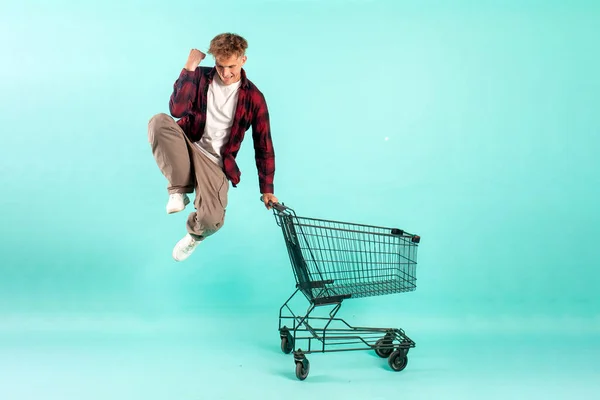young crazy guy shopper jumps and rejoices with shopping cart from supermarket on blue isolated background, male customer quickly runs in the air for shopping