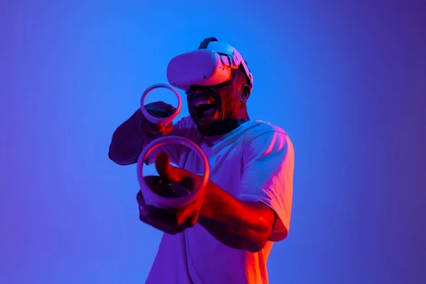 衝撃的なアフリカ系アメリカ人男性ゲーマーで仮想現実のメガネの撮影とネオンの照明でジョイスティックを目指して 男はガジェットとVrシューティングゲームをプレイし コントロールパネルと悲鳴を保持しています — ストック写真