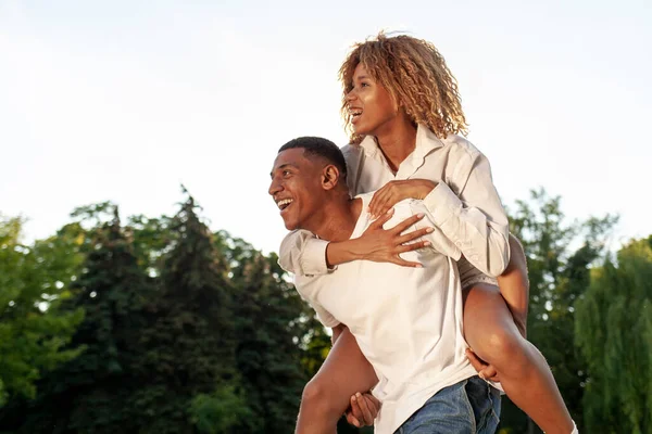 アフリカ系アメリカ人の幸せなカップルは夏に公園で一緒に歩いてコピースペースを見て 男は彼の背中に女性を運び 自由と関係の概念で前進します — ストック写真
