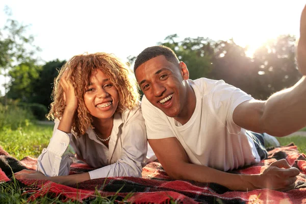 アフリカ系アメリカ人の若いカップルが草の上の夏に公園に横たわっており 自分自身を取り 男性と女性は自然の中でリラックスし ビデオリンクを介してオンラインで通信します — ストック写真