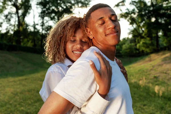 彼らの目を閉じて夢を見て公園に一緒に立って幸せな若いアフリカ系アメリカ人のカップル 彼女のボーイフレンドと笑顔を抱きしめる巻きガール 愛とケアの概念 — ストック写真