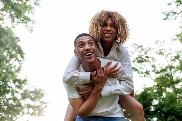アフリカ系アメリカ人の若いカップルが夏に公園で一緒に走り笑顔で男は彼のバック屋外で彼のガールフレンドを運びます 巻き毛の女の子抱擁男と先を見て — ストック写真
