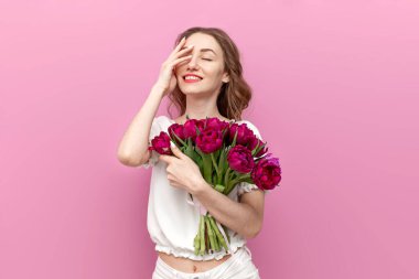 Şenlik kıyafetleri içinde bir buket çiçek tutan ve pembe arka planda gülümseyen, pembe laleli genç bir kız 8 Mart 'ı kutluyor.