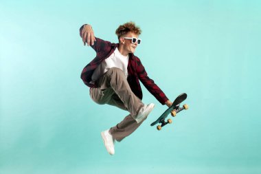 Genç çılgın adam kaykaya biniyor ve mavi izole arka planda zıplıyor, güneş gözlüklü hippi havada kaykayla uçuyor ve aşırı numaralar yapıyor.