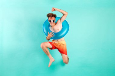 Şişirilebilir yüzme yüzüğüyle kazanan genç adam yazın mavi arka planda atlıyor ve uçuyor. Erkek turist plajda dinlenmek için koşuyor ve zaferle seviniyor.