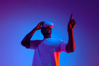 Sanal gerçeklik gözlüklü Afrikalı Amerikalı erkek oyuncu, neon ışıkta ellerine dokunuyor, modern VR cihazını kullanıyor ve fotokopi uzayını seçip işaret ediyor.