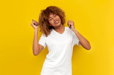 Sarı izole edilmiş arka planda dans eden genç kıvırcık Amerikalı kız gözleri kapalı hareket eden beyaz tişörtlü Afrikalı kadın.