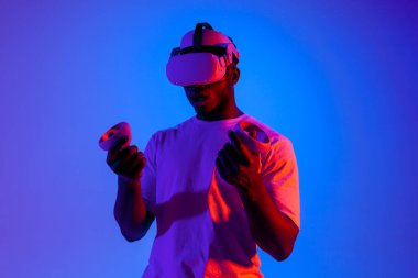 Sanal gerçeklik gözlüklü Afrikalı Amerikalı bir adam neon ışıkta joystickleri tutuyor, oyuncu gözlük kullanıyor ve kontrol panellerini tutuyor.