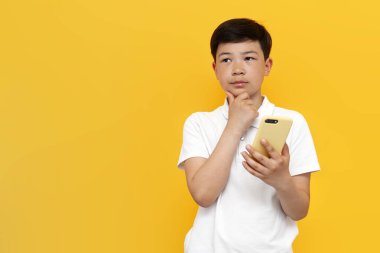 Beyaz tişörtlü, düşünceli Asyalı çocuk akıllı telefon kullanıyor ve sarı izole edilmiş arka plan üzerinde düşünüyor, Koreli çocuk tipleri cep telefonlarında ve rüyalarda online.