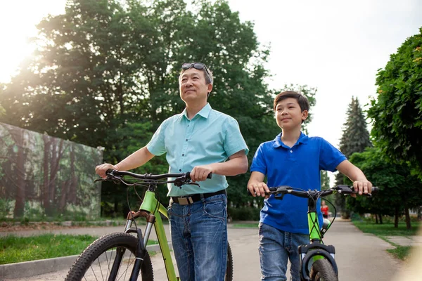 asian old father and son ride bikes together in the park, korean senior and grandpa are active with child in summer