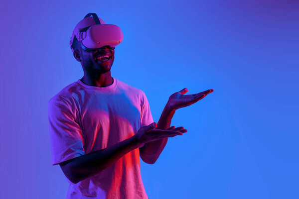 african american man in virtual reality glasses shows his hands to the side in neon lighting, the guy uses VR glasses and advertises copy space