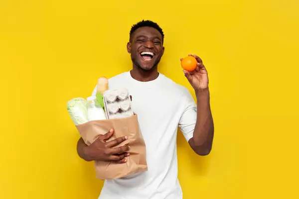 joyful male african american shopper holding eco bag with groceries and smiling on yellow isolated background, guy consumer holding shopping and eating