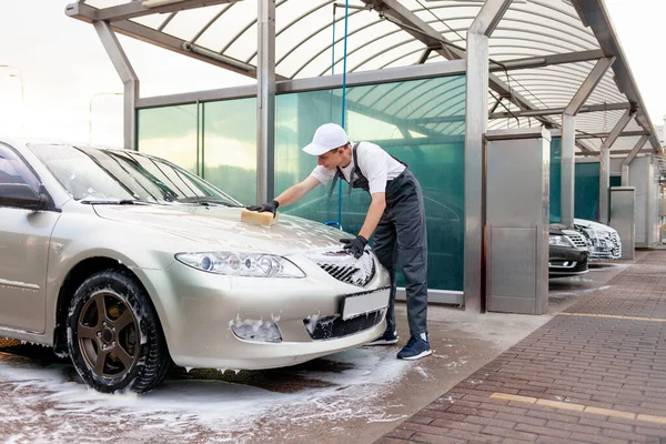 young guy in uniform car wash worker washes car with sponge with foam, man in overalls works in outdoors car cleaning service