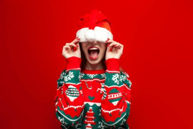 Noel süveteri içinde sevimli bir kız büyük bir Noel Baba şapkası içinde saklanıyor ve kırmızı arka planda çığlıklar atıyor. Utanmış bir kadın Noel kıyafetleri içinde gözlerini şapkayla kapatıyor ve ağzını açıyor.