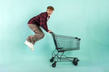 Genç çılgın adam alışveriş arabasıyla mavi izole edilmiş arka planda süpermarketten atlıyor ve seviniyor. Erkek müşteri hızla alışverişe koşuyor.