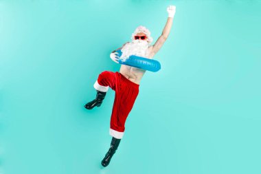 Takım elbiseli Noel Baba güneş gözlüklü şişme yüzme yüzüğü takıyor, zıplıyor, uçuyor ve mavi izole arka planda başarıyla seviniyor, Noel Baba denizde tatile çıkıyor ve zaferini kutluyor.