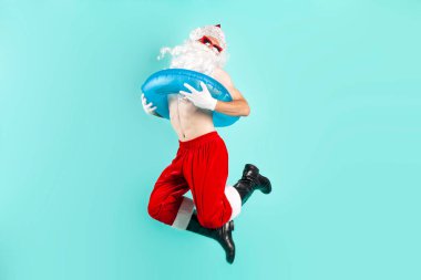 Takım elbiseli Noel Baba güneş gözlüklü şişme yüzme yüzüğü takıyor, zıplıyor, uçuyor ve mavi izole arka planda başarıyla seviniyor, Noel Baba denizde tatile çıkıyor ve zaferini kutluyor.