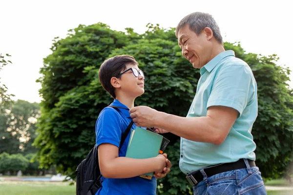 asian old father goes to school with son and carry books, korean boy with backpack and glasses goes to school with elderly dad in summer, grandfather take care of grandson
