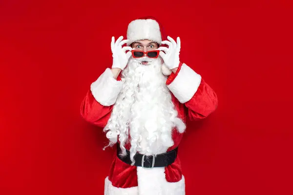 赤い眼鏡のショックを受けたサンタ クラスがカメラを見て驚き色の背景に驚きクリスマスのサンタの衣装を着た男 — ストック写真