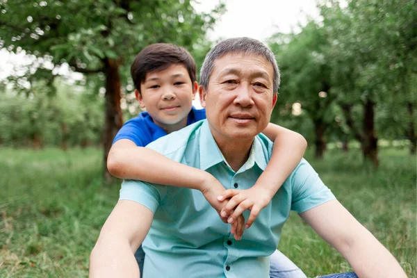 portrait of asian old father with little son outdoors, korean boy hugging grandpa and smiling in park, old man with child sit on grass