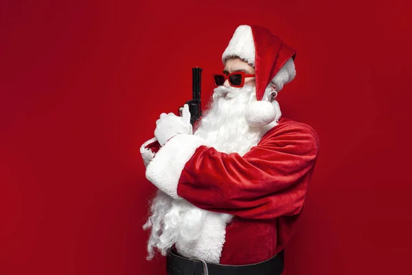 santa claus in hat and festive glasses holds gun in his hands on red background, dangerous man in santa costume with weapon threatens