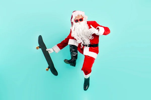 サングラスとスーツのサンタクロースは 青い隔離された背景にスケートボードでジャンプしてハエ ヒップスターサンタはスケートボードに乗り 新年とクリスマスのために空気をトリックします — ストック写真