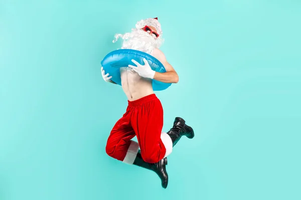 santa claus in suit with inflatable swim ring in sunglasses jumps and flies and rejoices in success on blue isolated background, santa tourist rides on vacation at the sea and celebrates victory