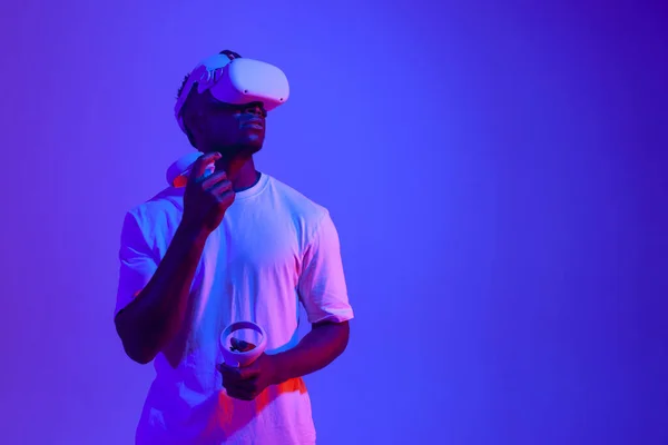 stock image african american male gamer in virtual reality glasses with joysticks looks away in neon lighting, guy plays with vr gadget and holds control panel and looks at copy space