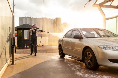 Üniformalı genç adam araba yıkama işçisi arabayı hortumla yıkıyor, tulumlu adam dışarıda araba temizleme servisinde çalışıyor.