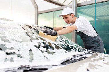 Üniformalı genç adam araba yıkama işçisi arabayı köpüklü süngerle yıkıyor, tulumlu adam dışarıda araba temizleme servisinde çalışıyor.