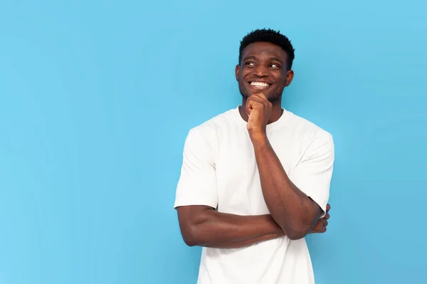 stock image pensive african american man in white t-shirt is dreaming and smiling on blue isolated background, the guy is planning and thinking and looking at the copy space