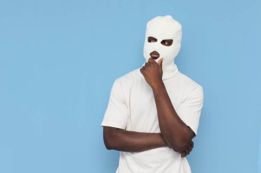 Beyaz kar maskeli Afro-Amerikan erkek kabadayısı mavi izole bir arka plan planlıyor ve düşünüyor. Maskeli adam hayal görüyor ve hayal görüyor.