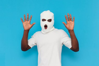 Ben pes ediyorum. Beyaz kar maskeli Afro-Amerikan erkek haydut mavi izole arka planda ellerini kaldırıyor, maske takmış zorba ağzı açık bir şekilde avuçlarını gösteriyor.
