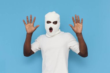Ben pes ediyorum. Beyaz kar maskeli Afro-Amerikan erkek haydut mavi izole arka planda ellerini kaldırıyor, maske takmış zorba ağzı açık bir şekilde avuçlarını gösteriyor.